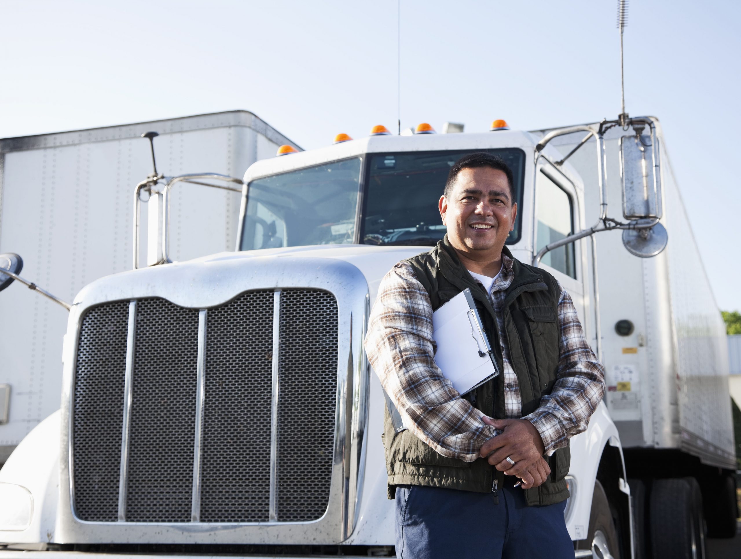 Truck driver (40s) standing in front of semi-truck with clipboard.